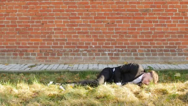 Un giovane uomo caucasico in un elegante abito nero si trova sull'erba e ride contro un muro di mattoni, slow-mo, spazio copia — Video Stock