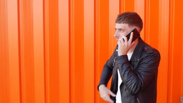 Unga orakad kaukasiska kille i en svart jacka som känslomässigt pratar i telefonen och är arg på den orange bakgrunden, långsamma-mo — Stockvideo
