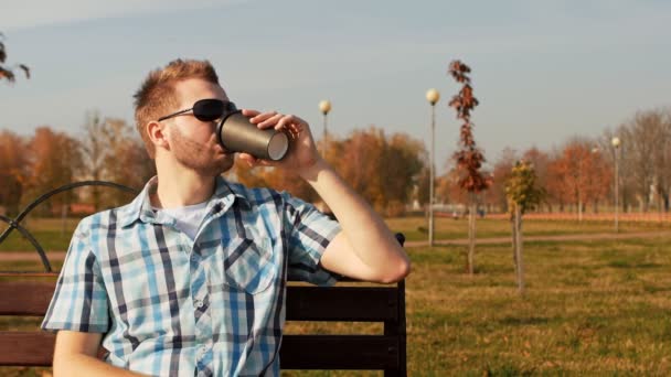 Tipo barbudo con estilo en gafas de sol se sienta en un banco y bebe café — Vídeo de stock