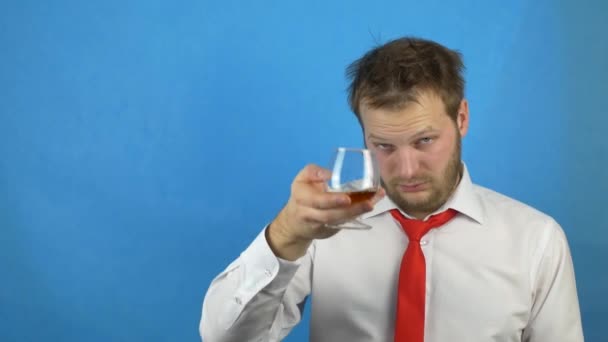 Hombre caucásico borracho con barba en camisa blanca y corbata bebiendo brandy de alcohol de un vaso, fondo azul, resaca — Vídeo de stock