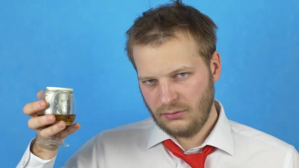 Молодой человек с бородой в белой рубашке и галстуке держит стакан алкоголя, в котором есть деньги, крупный план, алкоголь и пустая трата денег — стоковое видео