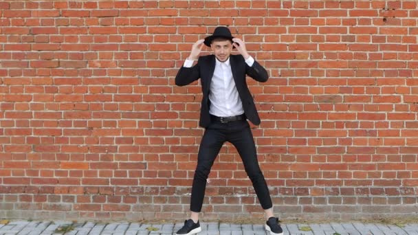 Jovem alegre homem dança contra uma parede de tijolo e tira o chapéu, caucasiano, câmera lenta, unbonnet, espaço de cópia — Vídeo de Stock