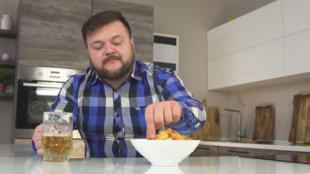 Uomo barbuto che beve birra e mangia patatine a casa in cucina, calorie, slow-mo — Video Stock