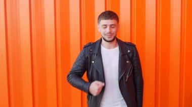 Esmer beyaz genç erkek siyah deri ceketli bir robot gibi hareket eder ve gibi gösterir bir turuncu arka plan, yakın çekim, slo-mo