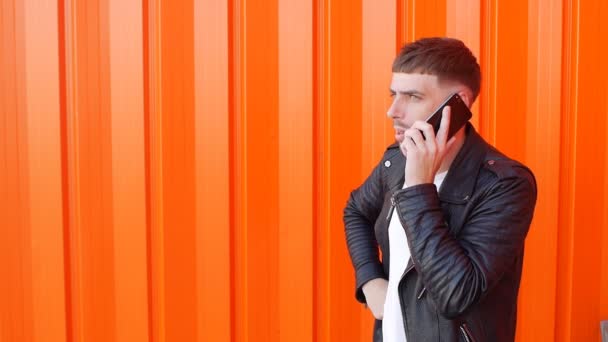 Jonge ongeschoren blanke man in een zwart jasje emotioneel praten aan de telefoon en is boos op de oranje achtergrond, slow-mo — Stockvideo