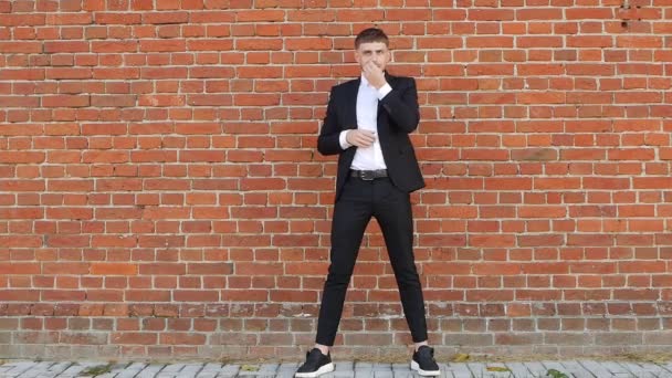 En ung kaukasiska man i en svart kostym står mot en tegelvägg och berör sin mustasch och skägg, långsamma-mo, kopia utrymme — Stockvideo