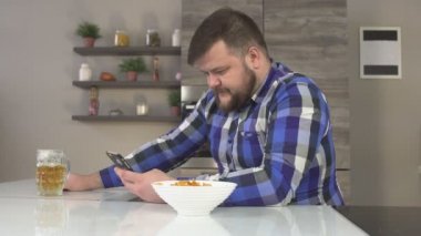 Sıkı beyaz adam mutfakta oturmuş, cips yeme ve içme bira, telefonu elinde ekose gömlekli