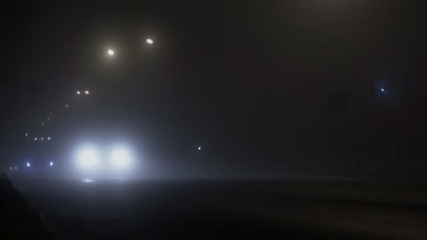 Транспортное средство с включенными фарами движется по дороге в тумане или тумане ночью, плохая видимость из-за тумана — стоковое видео