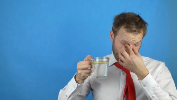 Молодой кавказский бизнесмен в белой рубашке и галстуке после похмелья пьет воду с лимоном, сонливостью, синим фоном, копировальным пространством — стоковое видео