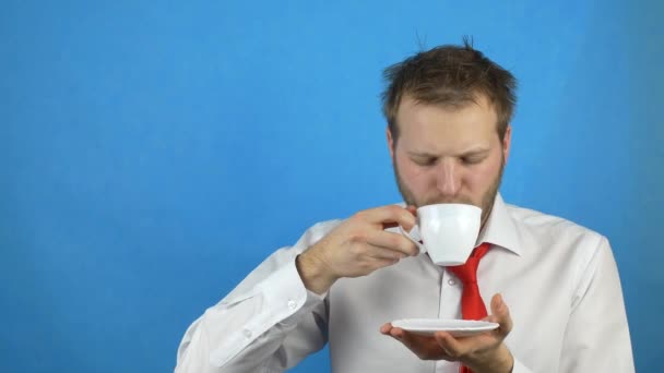 Junger Mann mit Bart, weißes Hemd und Krawatte, Kaffee trinkend mit Kater und Schläfrigkeit, blauer Hintergrund, Kopierraum — Stockvideo