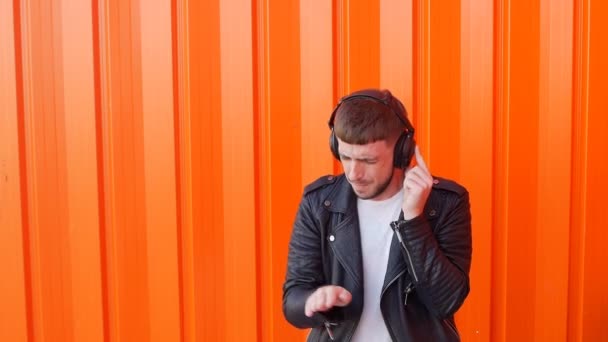 Молодой кавказский мужчина в музыкальных наушниках слушает музыку и танцы на оранжевом фоне, скачет, замедленное движение, закрылки для ушей — стоковое видео