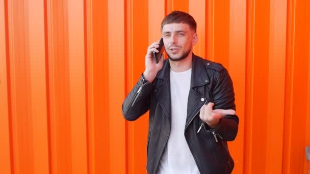 Young unshaven caucasian man in a black jacket talking on the phone on an orange background, slow-mo, fashionable — Stock Video