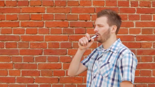 Fashionable and stylish guy with a beard smoking a vape against a brick wall, copy space, slow-mo, outside — Stock Video