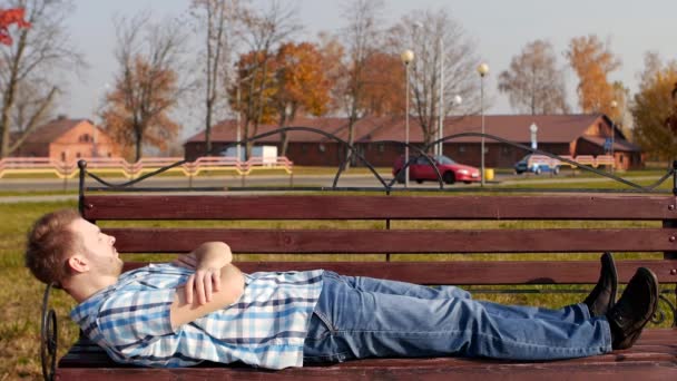 Joven guapo y elegante hombre caucásico se encuentra y descansa en un banco en el parque, espacio de copia, cámara lenta — Vídeos de Stock