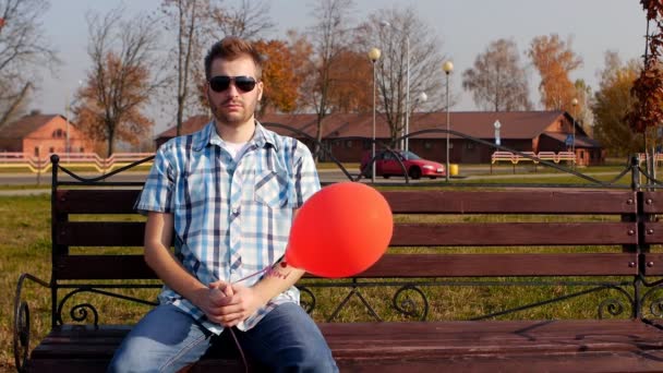 Brutale jonge man in zwarte bril zit op een bankje met een rode ballon en kijkt naar de camera, kopie ruimte, slow motion, lifestyle — Stockvideo