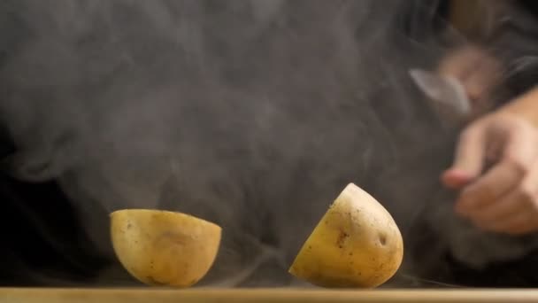 Un homme coupe avec un couteau une pomme de terre fraîche dans la brume et l'évaporation, gros plan, fond noir, slow mo, végétarien — Video