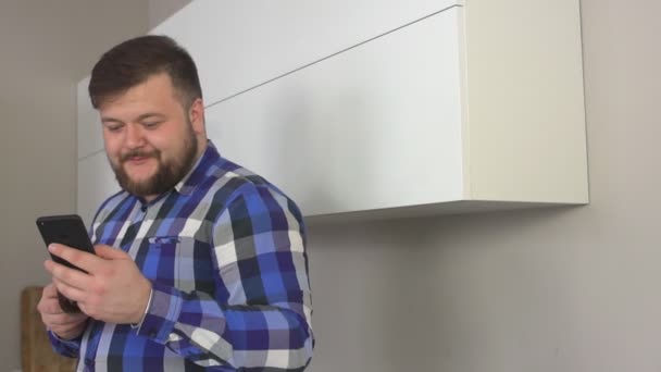 Uomo caucasico soddisfatto con la barba che beve caffè in cucina e guarda il telegiornale al telefono, primo piano, rallentatore — Video Stock