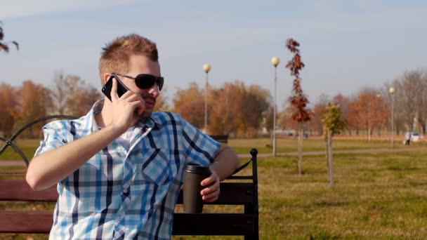 Giovane uomo elegante e alla moda in occhiali neri si siede su una panchina a bere caffè e parlare al telefono, primo piano, spazio copia, rallentatore, alla moda — Video Stock