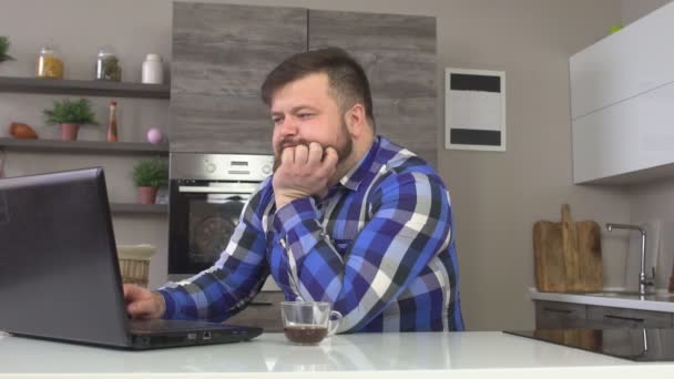 Un homme avec une barbe est assis dans la cuisine avec un ordinateur portable et boire le café du matin, ralenti mo — Video