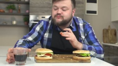 Şişman beyaz adam içinde belgili tanımlık mutfak yiyor lokanta hamburger ve çizburger oturur ve zararlı soda, yavaş hareket ile limonata içer