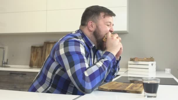 Full kaukasiska man med ett skägg som äter en hamburgare i köket, snabbmat och tvättar ner med saft, långsamma-mo — Stockvideo