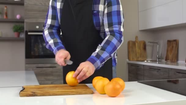 Een man in een schort snijdt een sinaasappel in de keuken, vitamine, close-up, langzame mo, achtergrond — Stockvideo