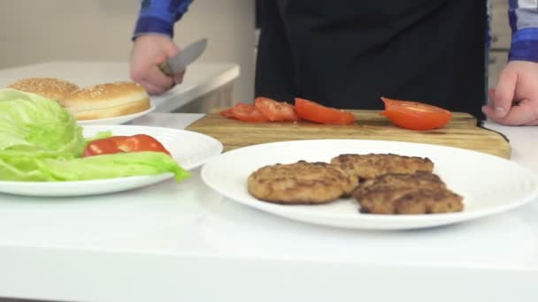 Egy férfi egy kötény a konyhában készül egy burger, az asztalon ezek a hozzávalók egy sajtburgert és szeletelt paradicsomot, lassú mozgás — Stock videók