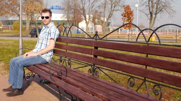 Young handsome and fashionable caucasian guy in black glasses sits on a bench and looks into the camera, slow-mo, copy space, outside — Stock Video