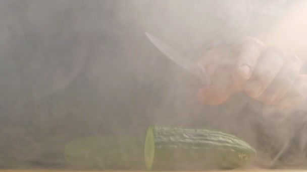 Un hombre corta un pepino jugoso granjero con un cuchillo en neblina y evaporación, fondo negro, primer plano, vegetal, cámara lenta — Vídeo de stock