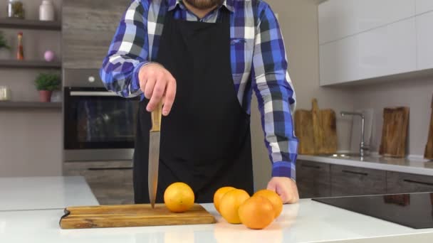 Um homem em um avental em uma cozinha moderna com uma faca, laranjas estão sobre a mesa, close-up, câmera lenta, frescura — Vídeo de Stock