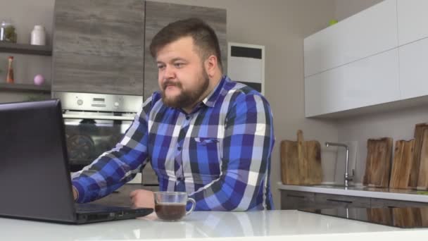 Un homme caucasien sérieux et concentré boit du café et regarde dans un ordinateur portable, travaille à la maison, cuisine, ralenti, ordinateur — Video