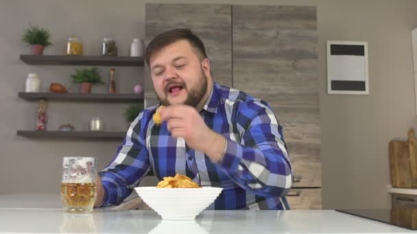 Мужчина с бородой на клетчатой рубашке сидит на кухне и пьет пиво с чипсами, удовольствие от еды, медлительность — стоковое видео