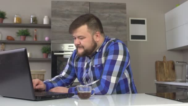 Un hombre con barba está sentado en la cocina, bebiendo café y mirando el portátil con decepción, perdiendo, cámara lenta, negocios — Vídeos de Stock