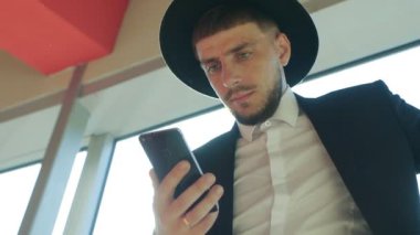 Bir çanta ve şapkası sakallı şık beyaz erkek işadamı duran ve bir akıllı telefon İşletme Merkezi, yakın çekim, Güneş, slo-mo, banka bilgileri okuma