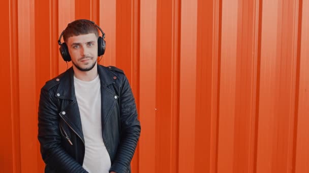 Portrait of young fashionable positive man in earphones or headphones near orange wall background — Stock Video