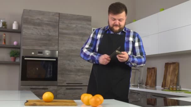 Un hombre gordo con barba está afilando un cuchillo en una cocina moderna, preparándose antes de cocinar en la cocina, cámara lenta, frescura — Vídeos de Stock