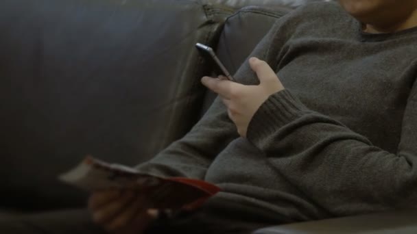 Мужчина с брошюрой или буклетом сидит на диване и пользуется смартфоном — стоковое видео