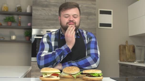 Un hombre gordo con barba se lame los dientes y quiere comer hamburguesas con queso, alegría, deseo de comida, exceso de peso, mes lento, hamburguesa — Vídeo de stock