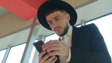 Bir çanta ve şapkası sakallı şık beyaz erkek işadamı duran ve iş merkezi, yakın çekim, Güneş, ağır çekim bir akıllı telefon bilgileri okuma
