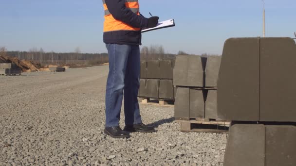 Un inspector con un chaleco de señalización y un casco blanco con gafas negras toma notas en su carpeta sobre la calidad de la acera y la construcción de carreteras — Vídeos de Stock