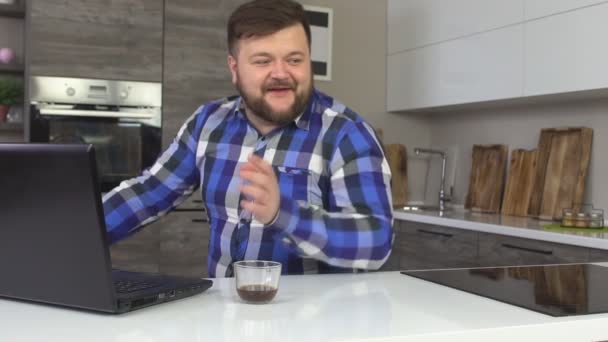 Um homem contente e alegre com barba trabalha em um laptop em uma cozinha moderna e ri, câmera lenta, tecnologia — Vídeo de Stock