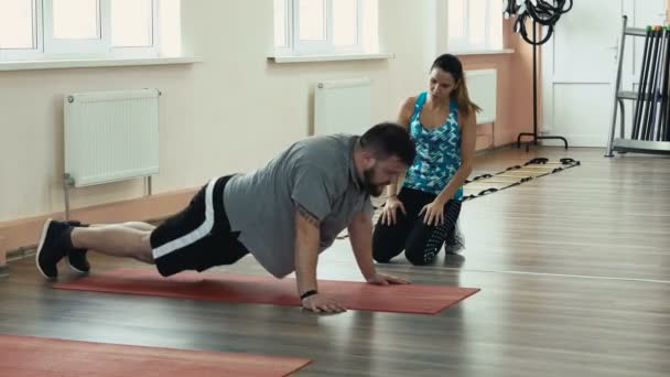 Thick obese guy together with personal slim coach do physical jerks in gym. Overweight male and female trainer with perfect body burn calories by workout. Personal training from athletic girl for — Stock Video