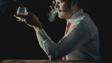 Sakallı adam kadeh alkollü içki ya da içecek ile oturur ve sigara içiyor