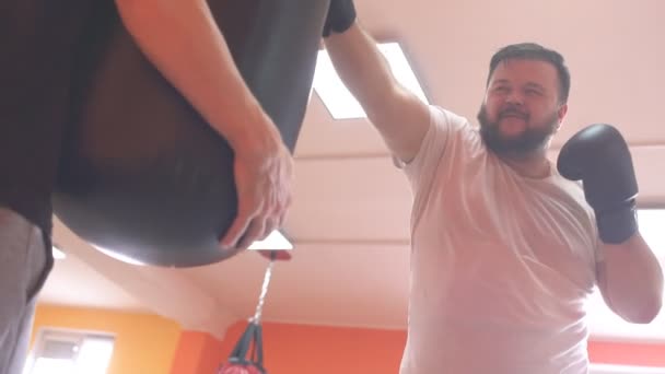 Толстый бородач боксирует и бьет боксерскую грушу боксёрскими перчатками, замедленным движением — стоковое видео