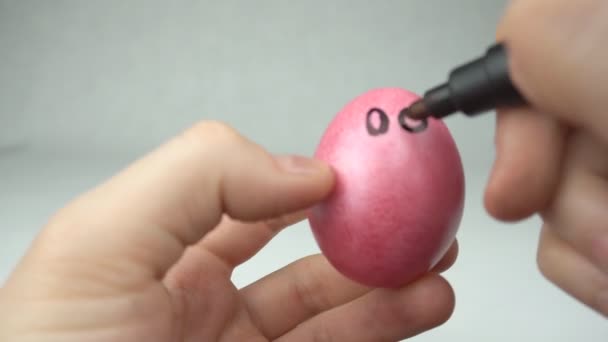 Ένας άνθρωπος χρωματίζει με μια αισθητή-πένα tip ένα κόκκινο Πασχαλινό αυγό αντλεί ένα smiley και ένα πρόσωπο, μια αργία, γκρο πλαν — Αρχείο Βίντεο