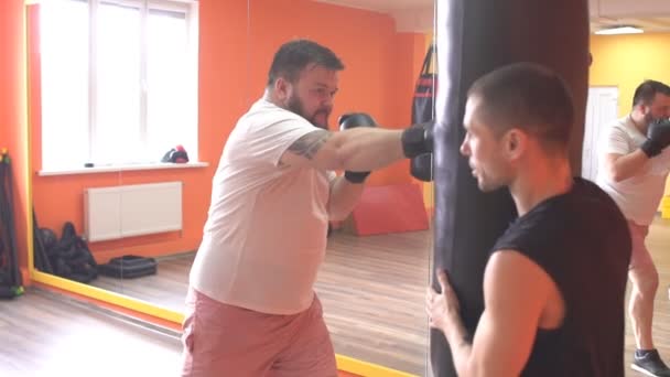 Hombre gordo barbudo cumple un golpe en el boxeo, golpea un saco de boxeo bajo la supervisión de un entrenador, cámara lenta — Vídeo de stock