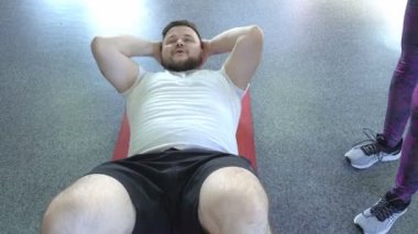 Sakallı şişman beyaz bir adam, bir fitness eğitmeni gözetiminde karın için egzersizler yapar, yakın çekim