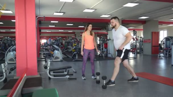 Grasso uomo caucasico con la barba in palestra esegue un esercizio squat con manubri sotto la supervisione di una bella ragazze personal trainer, accovacciato — Video Stock