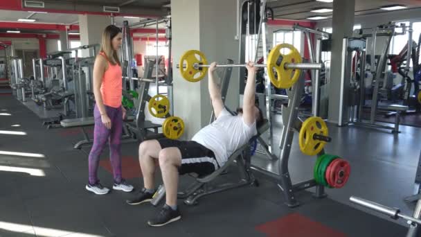 Gordo caucásico hombre con barba realiza un ejercicio de press de banca dumbbell en su espalda mientras asegura una hermosa chica entrenador — Vídeo de stock