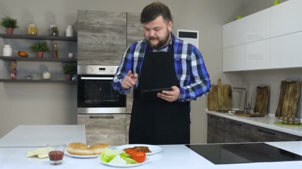 Grueso barbudo hombre comprueba o verifica los ingredientes de una hamburguesa en una cocina — Vídeos de Stock